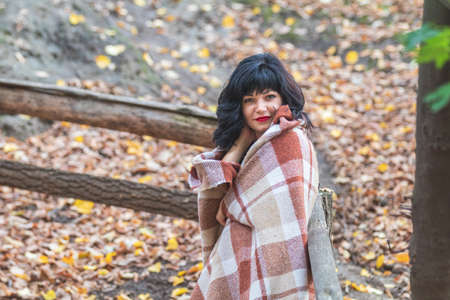Cute happy beautiful sexy brunette female girl resting sitting in autumn garden on the steps, wrapped in a woolen plaid blanket.の写真素材