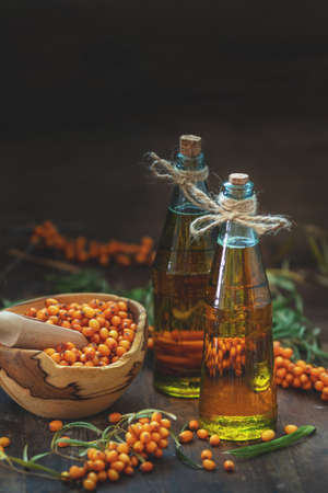 Natural, organic sea-buckthorn berry in bowl and sea buckthorn oil in glass vintage bottle on dark wooden background. Dark rustic style, natural remedy.の写真素材