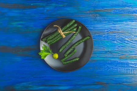 Kabanosy, sausages green with wasabi made of pork in plate on a dark blue wood surface with addition of fresh herbs, chili pepper and spicesの写真素材