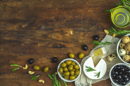 Set of cheese camembert, black and green olives, quail eggs on plates, olive oil and rosemary, cashew nuts, on old dark wooden table background. Top view, copy space.