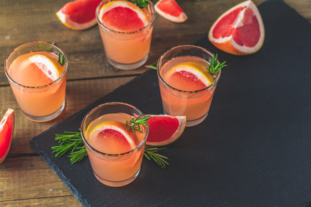 Fresh pink alcoholic cocktail with grapefruit, ice and rosemary, drink glass on a black stone board,...