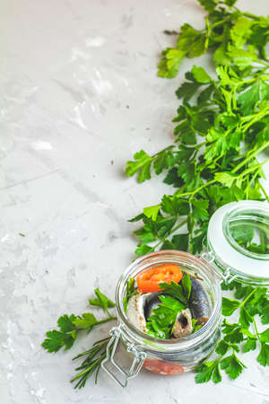 Sardines or baltic herring with rosemary, thyme, parsley, tomatoes slices and spaces in glass jar on light gray concrete table surface. Preserved marinated sea fish, copy space for you text.の写真素材