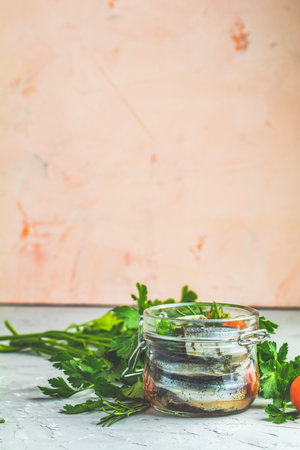 Sardines or baltic herring with rosemary, thyme, parsley, tomatoes slices and spaces in glass jar on light gray concrete table surface. Preserved marinated sea fish, copy space for you text.の写真素材