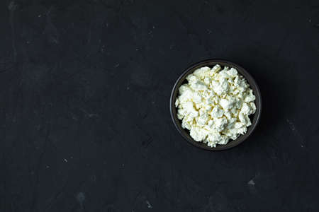 Homemade cottage cheese in a black ceramic bowl on dark concrete table surface, top view, copy space.の写真素材