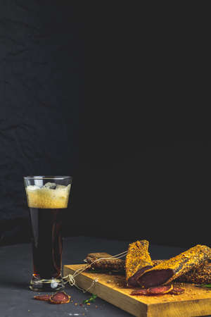 Dark beer in glass and Jerky, basturma, dried meat beef, meat smoked jerky with spices on wooden cutting board, black concrete surface table background.の写真素材
