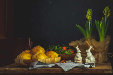 Happy Easter greeting card with homemade buns in wooden plate standing with crocuses flowers and bunny decor on linen tablecloth. Copy space, dark rustic style.の写真素材