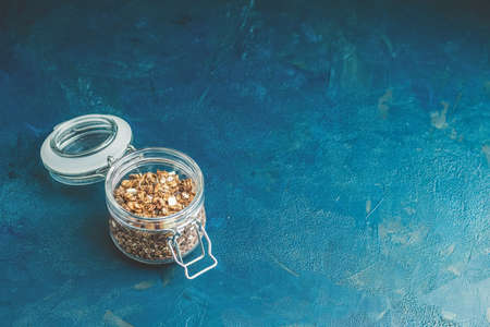Open glass jar of organic granola with berries, coconut chips and seeds on a dark blue concrete table surface.の写真素材