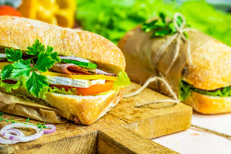 Italian porchetta sandwich. Ciabatta sandwich with ham, tomato, cheese, pepper, onion and salad on wooden cutting board with ingredients.の写真素材