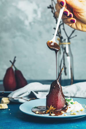 Chocolate sauce pours from a spoon on red pears in wine. Red pears are in a blue plate. The concept of still life on blue concrete surface.の写真素材