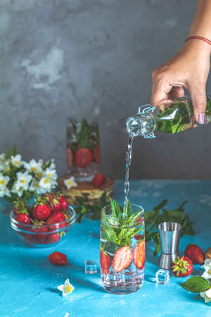Woman pouring lemonade from bottle into glass. Detox infused water with strawberry and mint in highball glasses on blue concrete table background, copy space. Cold summer drink. Mineral water.の写真素材