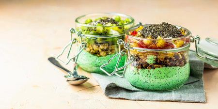 Two chia seed pudding with matcha green tea, kiwi and granola, orange in glasses on light pink concrete background. Healthy breakfast. Banner with copy space. Healthy eating conceptの写真素材