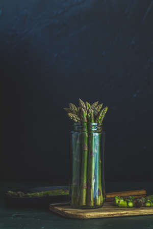 Green asparagus in glass jar standing on table at dark wall background. Fresh asparagus store. Spring seasonal vegetables. Copy spaceの写真素材
