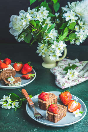 Chocolate rollcake with fresh strawberries in ceramic plate, jasmine and white peonies on dark green concrete surface tableの写真素材