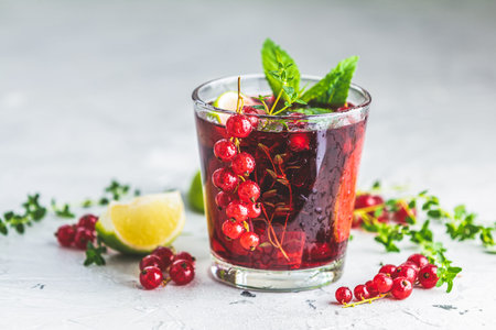 Cold red cocktail with currant, lime, mint, thyme and ice in glass on light gray concrete surface...