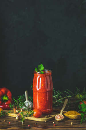 Homemade DIY natural canned hot tomato sauce chutney with chilli or adjika in glass jar standing on wooden table with cherry tomatoes, salt, pepper,  herbs, selective focusの写真素材