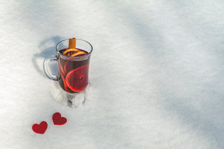 Cup with delicious mulled wine and spices on a snow covered background decorated with heart. Beautiful sunny winter day, Valentines Day greeting card with copy space for you text.の写真素材