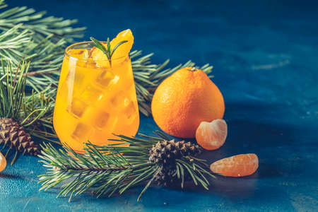 Yellow orange cocktail with tangerine and rosemary in glass on dark blue concrete background decorated pine branches with cones, close up. Christmas and New Year holiday welcome drinkの写真素材