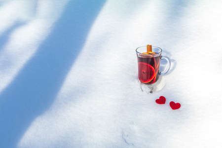 Cup with delicious mulled wine and spices on a snow covered background decorated with heart. Beautiful sunny winter day, Valentines Day greeting card with copy space for you text.の写真素材