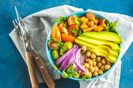 Healthy food. Healthy vegan lunch bowl of fried sweet potato, chickpeas, red onion, avocado, tomatoes, and brussels sprouts. Top view, classic blue concrete surface, flat lay.の写真素材
