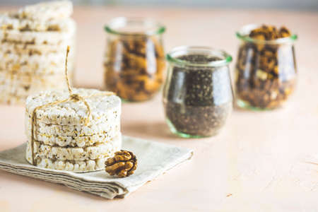 American puffed rice cakes. Healthy snacks with almonds, raisins, peanuts, pistachios in glass jars on light pink concrete surface.の写真素材