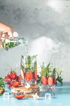 Women hand is pouring detox water from glass bottle to glass with strawberries, ice and mint. Summer...