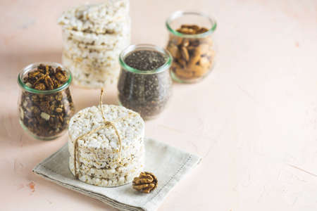 American puffed rice cakes. Healthy snacks with almonds, raisins, peanuts, pistachios in glass jars on light pink concrete surface. Copy space for you text.の写真素材