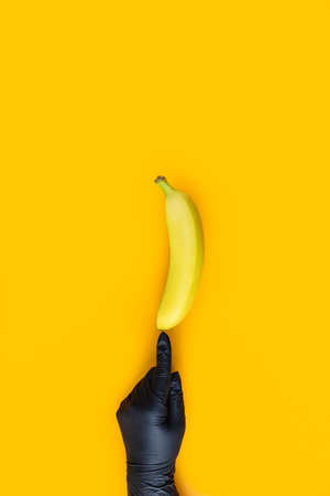 Hand in black glove holds banana on yellow background. Food art. Conception coronavirus new lifestyle concept.の写真素材