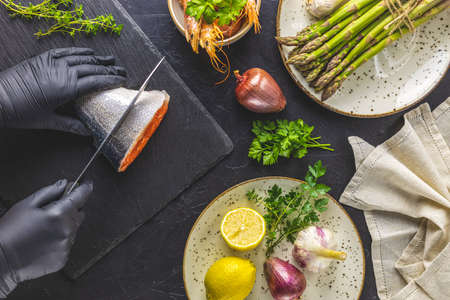 Hands in black gloves cut trout fish on black stone cutting board surrounded herbs, onion, garlic, asparagus, shrimp, prawn in ceramic plate. Black concrete table surface. Healthy seafood background.の写真素材
