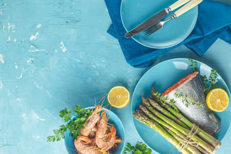 Fresh raw asparagus with trout fish, lemon and parsley, shrimp, prawn  in blue ceramic plates with napkin on light blue concrete table surface. Healthy food background with copy space for you text.の写真素材