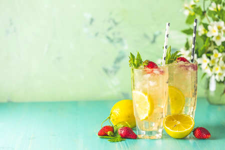 Two glasses of cold icy refreshing drink with lemon and strawberry served with bar tools on green wooden table with white blooming flowers. Fresh cocktail drinks with ice fruit and herb decoration.の写真素材