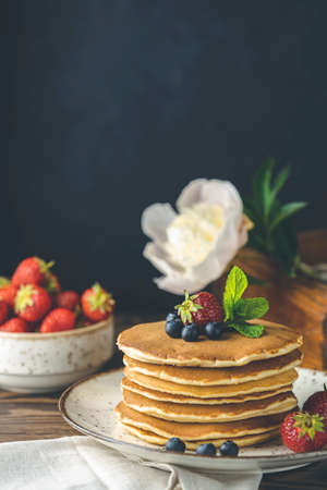 Pancake with srtawberry, blueberry and mint in ceramic dish, syrup from small ceramic jar and flowers on a dark wooden table and black background. の写真素材