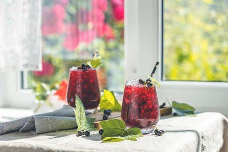 Two glass of cold ice black currant juice with ripe berries and green leaves on table in sunny room near window with garden outside.の写真素材