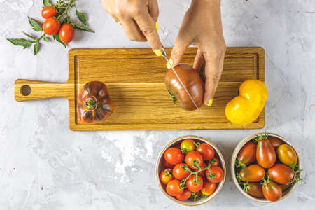 Woman hands cut the tomato on wooden board. Colorful tomatoes in ceramic plates on light gray concrete surface. Concept of green house life and products of subsistence farming, flat lay, copy spaceの写真素材