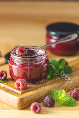 Two glass jar of fresh homemade raspberry jam on a wooden background. Several fresh berries and mint...