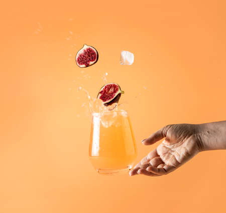 Woman hand support fly glass of pink cocktail drink with splash, juice slice fig falling in glass. Summer art food concept on peach color backgroundの写真素材