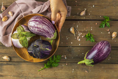 Woman hand hold wooden pot with different raw fresh eggplant. Dark wooden background. Concept of green house life style and products of subsistence farming, flat lay, copy spaceの写真素材