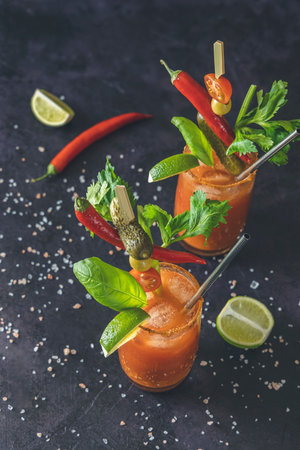 Two glasses of tomato juice with vodka, celery, spices, salt and ice in portion glasses with copy space. Bloody Mary cocktail. Alcoholic drink and ingredients at dark concrete table surface.の写真素材