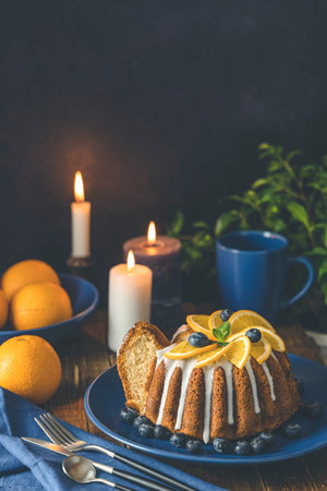 Orange bundt cake with blueberry surrounded fruits, candles, plant and cutleries on dark wooden...