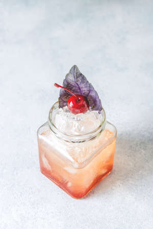 Picnic smash. Refreshing cold summer cocktail with grapefruit and  garnish by violet red basil leaves and cherry. Served in glass jar with crashed ice. Light gray table surface.の写真素材