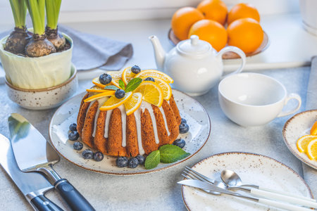 Orange bundt cake with blueberry surrounded fruits, plant and cutlery on light table near window....