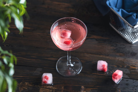 Elegant glass of pink rose champagne with frozen raspberries in ice cube and water drops on dark...