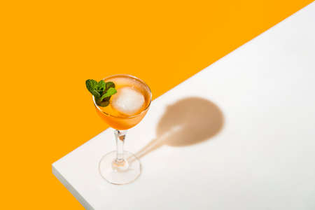 Elegant stemware glass of fresh orange cocktail with big ice ball on white table surface, bright yellow background. Spring summer art drink food concept.の写真素材