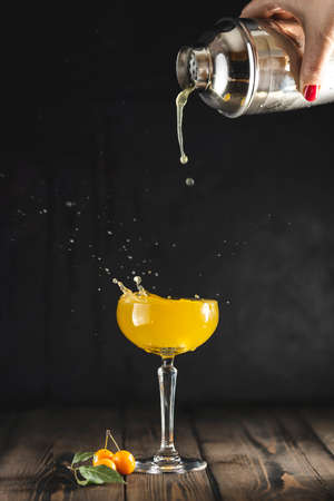 Woman hand pouring plum gin and tonic cocktail from shaker in champagne coupe glass with splash. Dark wooden table surface.の写真素材