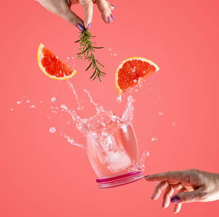 Woman hand support fly glass of grapefruit drink with splash, juice grapefruit slices falling in glass. Cocktail of grapefruit, thyme and rosemary flavor. Summer art food concept on pink backgroundの写真素材