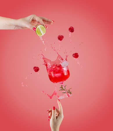 Woman hand support fly glass of raspberry drink with splash, juice raspberry falling in glass. Cocktail of raspberry, lime and rosemary flavor. Summer art food concept on pink backgroundの写真素材