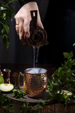 Woman hand pouring Green Apple Irish Mule cocktail cocktail from shaker in copper mug surrounded by ingredients and bar tools on dark wooden table surfaceの写真素材