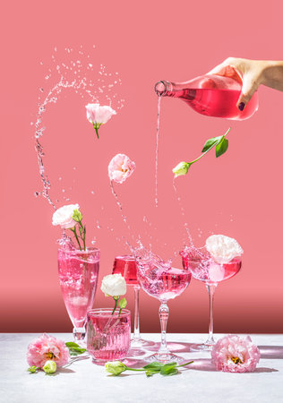 Different elegant champagne glasses with sparkling wine, women hand is pouring rose wine from bottle to coupe glass. Splash, splatter, fly flowers with water drops on the pink background. Art food or drink concept.の写真素材
