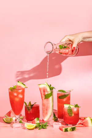 Different elegant glasses with watermelon cocktail, women hand is pouring sparkling water from bottle to glass. Splash, splatter, water drops on the pink background. Art food or drink concept.の写真素材