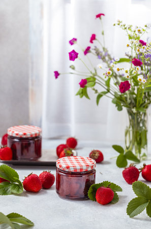 Fresh homemade strawberry jam in glass jar on a light background. Several fresh berries, green strawberries leaf and flowers are near it.の写真素材