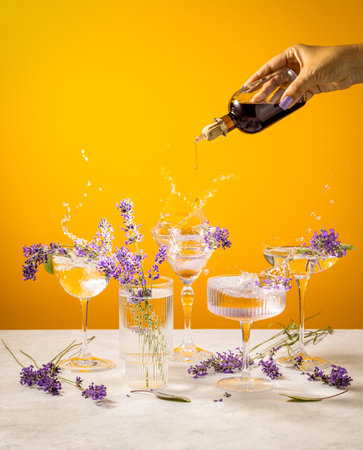 Hand pouring syrup from a bottle into a cocktail glass decorated with lavender flowers on a yellow backgroundの写真素材